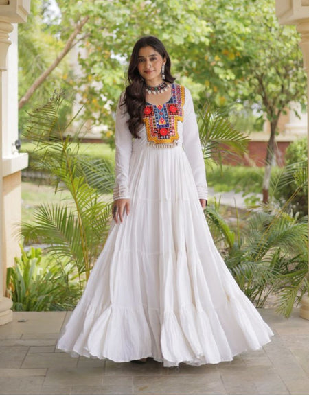 A woman wearing a long white maxi dress with colorful embroidered detailing on the chest stands in a garden corridor framed by beige pillars.