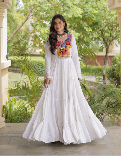 Load image into Gallery viewer, A woman wearing a long white maxi dress with colorful embroidered detailing on the chest stands in a garden corridor framed by beige pillars.
