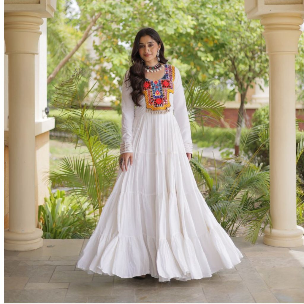 A woman wearing a long white maxi dress with colorful embroidered detailing on the chest stands in a garden corridor framed by beige pillars.