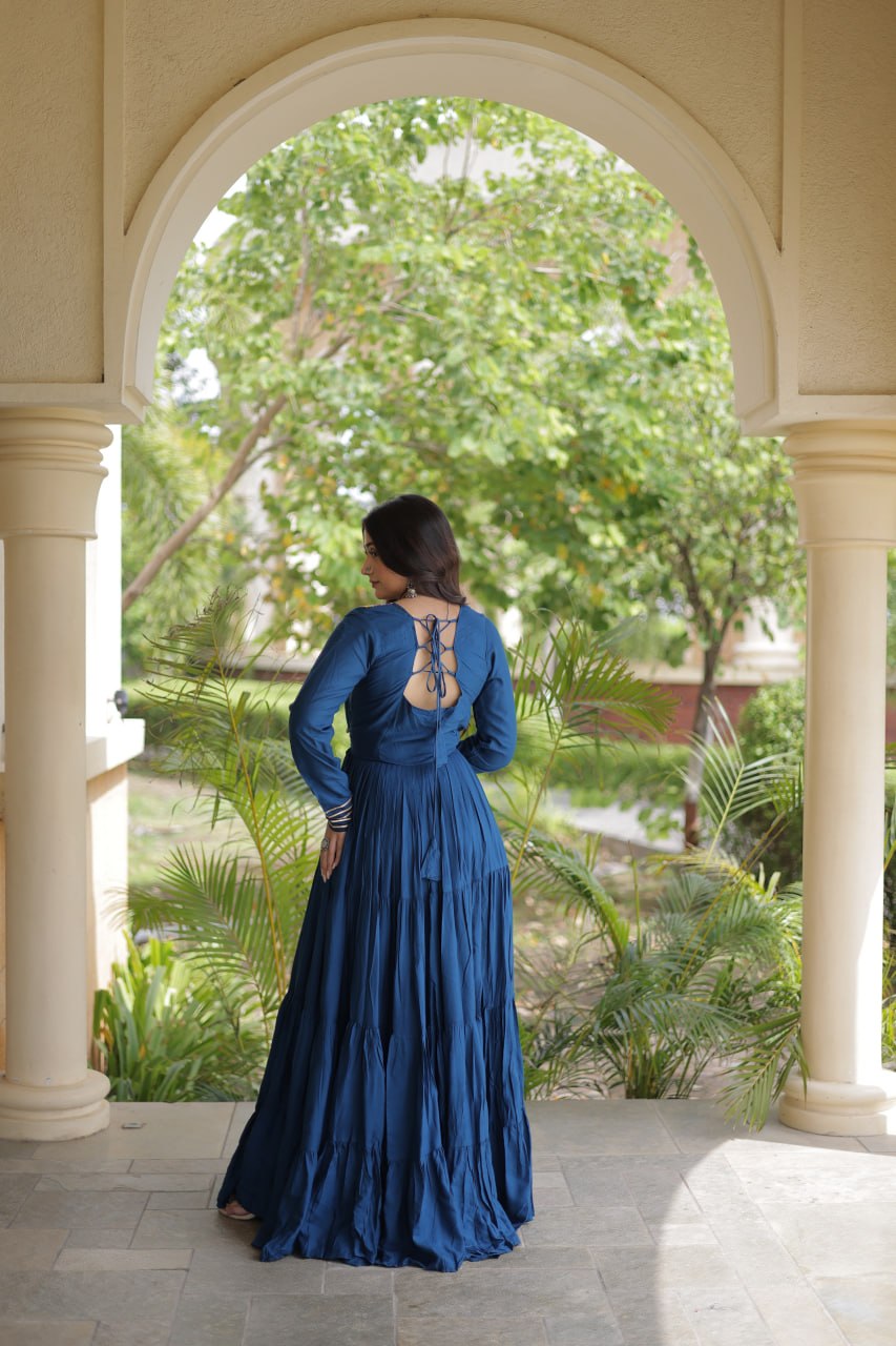 A woman wearing a long-sleeved navy blue maxi dress with an open lace-up back, standing in a stone archway with a lush garden background.