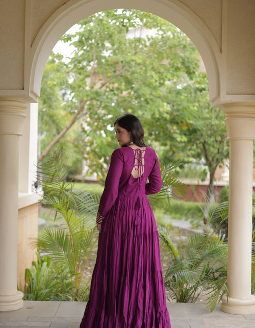 Load image into Gallery viewer, A woman wearing a long, pleated magenta-purple maxi dress with a lace-up open back, standing in a stone archway with a lush green garden background.
