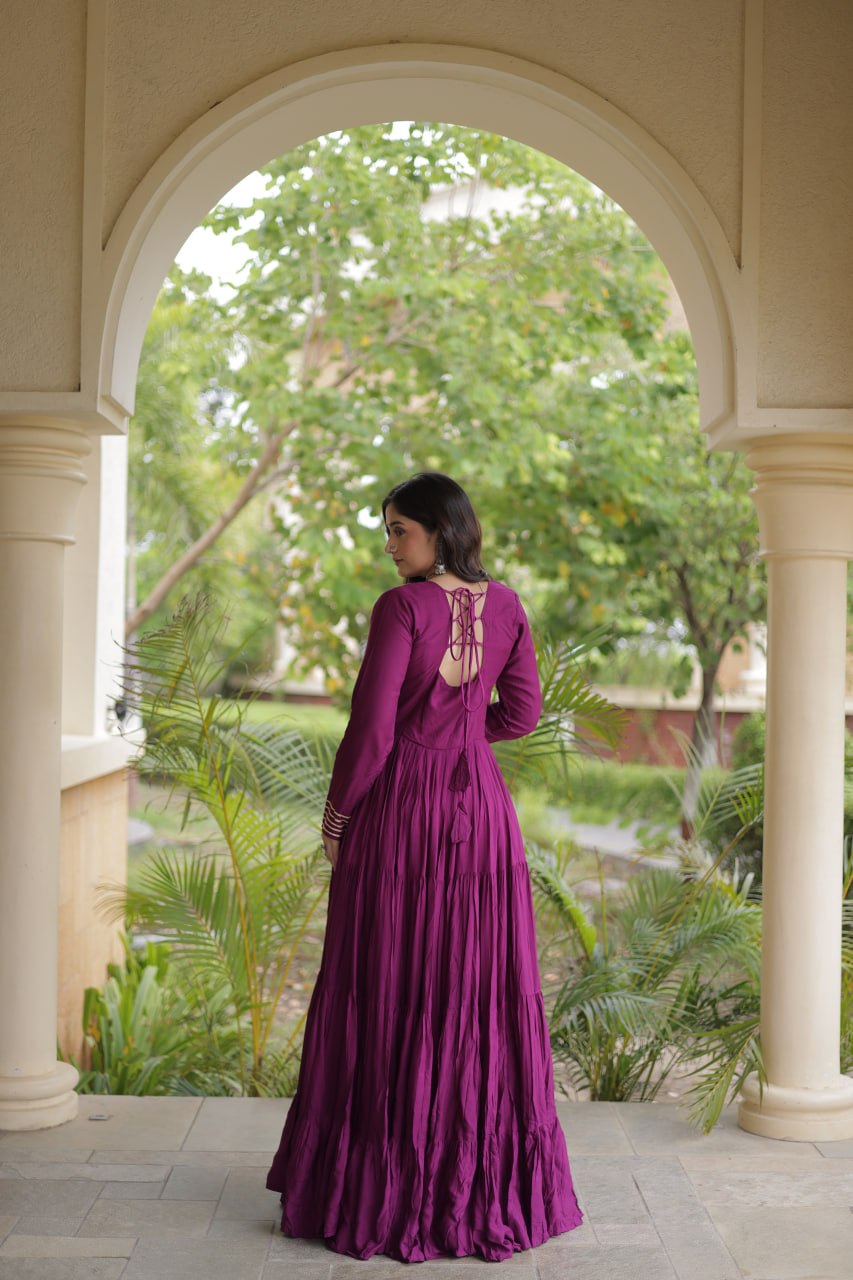 A woman wearing a long, pleated magenta-purple maxi dress with a lace-up open back, standing in a stone archway with a lush green garden background.
