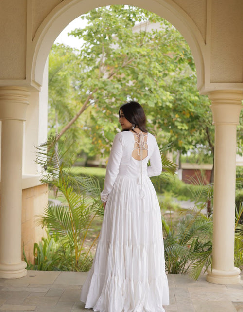 Load image into Gallery viewer, A woman wearing a long-sleeved white tiered wedding dress with an open back and lace-up detail, standing in a stone colonnade with green garden foliage visible through the archway.
