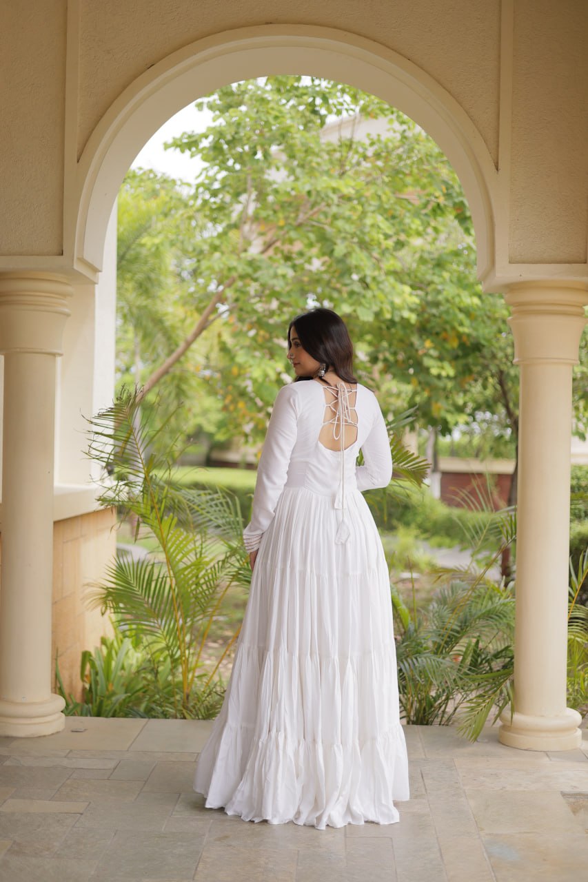 A woman wearing a long-sleeved white tiered wedding dress with an open back and lace-up detail, standing in a stone colonnade with green garden foliage visible through the archway.