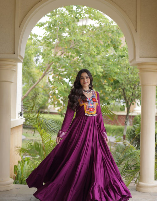 Load image into Gallery viewer, A woman wearing a long-sleeved magenta purple Anarkali gown with a colorful embroidered chest panel, standing in a courtyard framed by a large archway.
