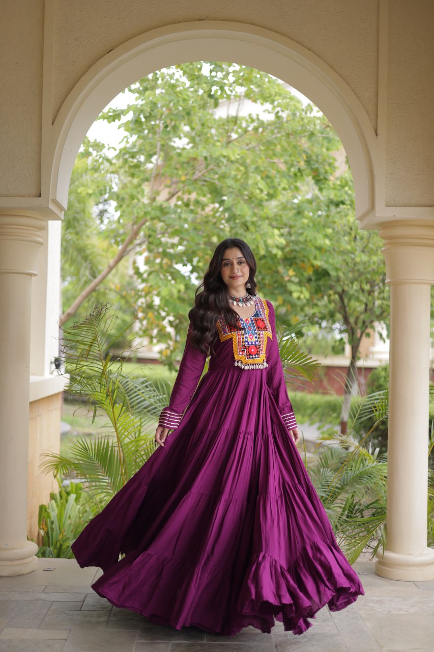 A woman wearing a long-sleeved magenta purple Anarkali gown with a colorful embroidered chest panel, standing in a courtyard framed by a large archway.