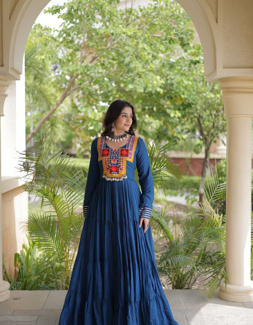 Load image into Gallery viewer, A woman wearing a long-sleeved navy blue maxi dress with colorful embroidered detailing on the chest stands under a beige stone archway with green trees in the background.
