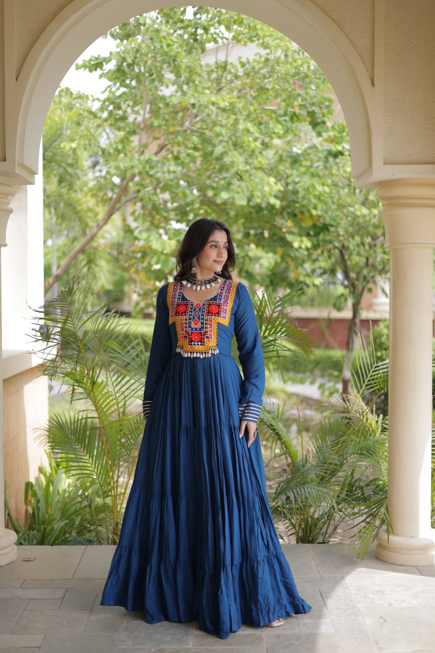A woman wearing a long-sleeved navy blue maxi dress with colorful embroidered detailing on the chest stands under a beige stone archway with green trees in the background.
