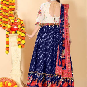 A woman wearing a navy blue lehenga choli with white floral prints, a white embroidered blouse, and a red and pink patterned dupatta, standing in front of a beige wall decorated with marigold garlands.