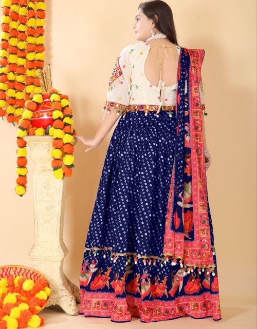 Load image into Gallery viewer, A woman wearing a navy blue lehenga choli with white floral prints, a white embroidered blouse, and a red and pink patterned dupatta, standing in front of a beige wall decorated with marigold garlands.
