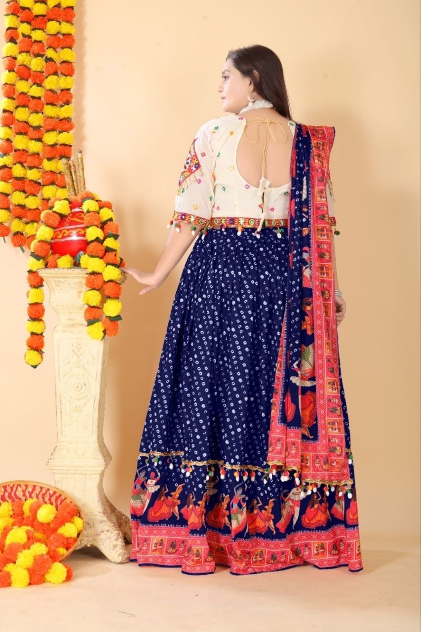 A woman wearing a navy blue lehenga choli with white floral prints, a white embroidered blouse, and a red and pink patterned dupatta, standing in front of a beige wall decorated with marigold garlands.