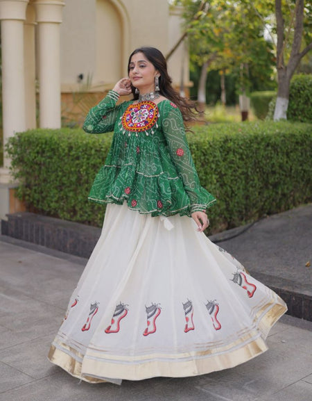 A woman wearing a green embroidered kurta with a long white embroidered skirt, standing outdoors in front of a garden and building.