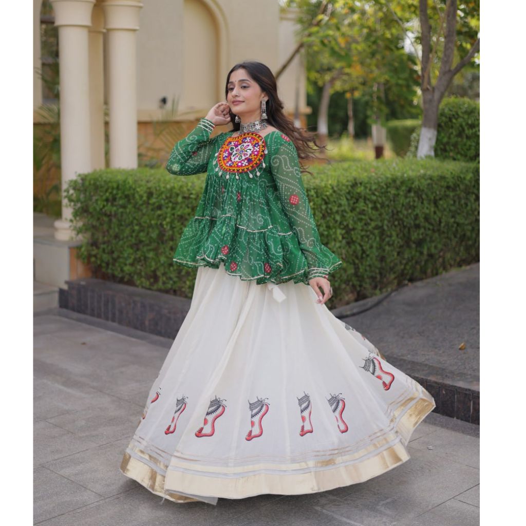 A woman wearing a green embroidered kurta with a long white embroidered skirt, standing outdoors in front of a garden and building.