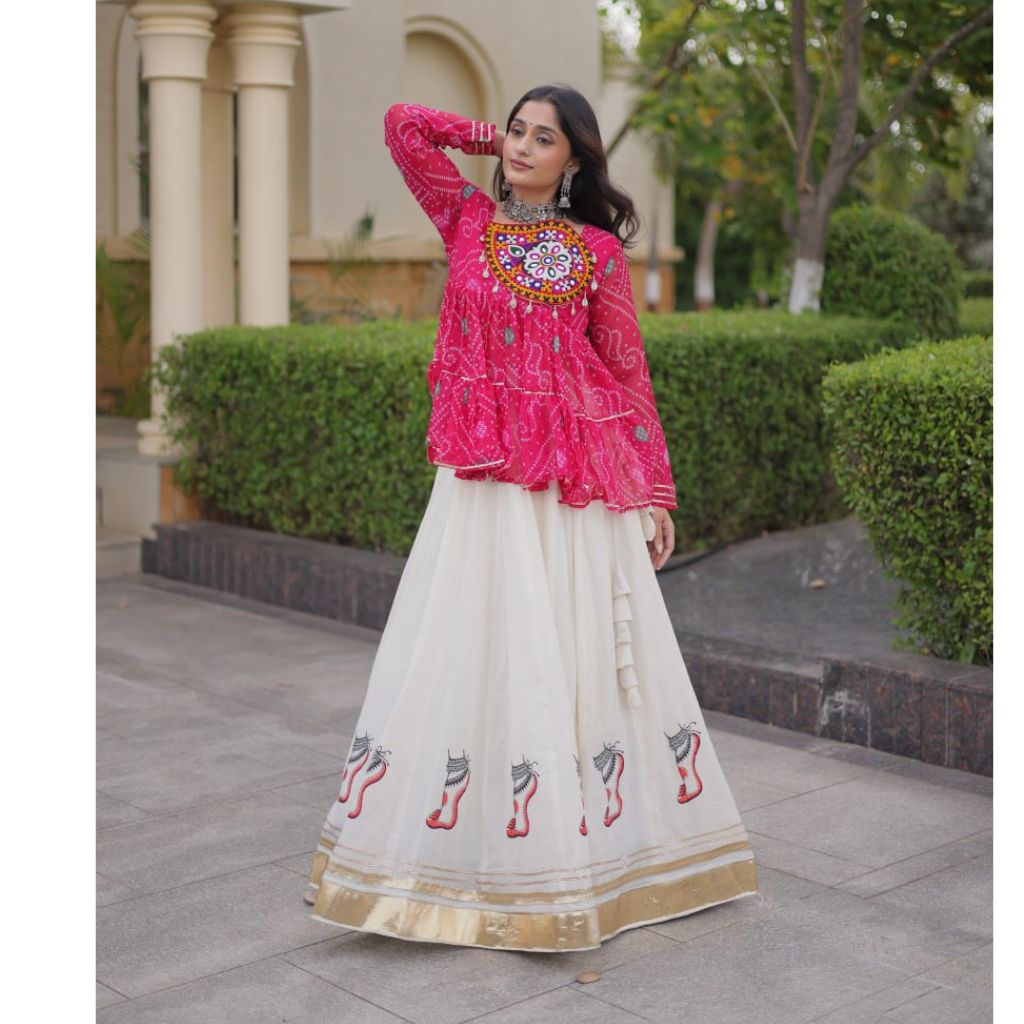 A woman modeling a traditional Indian outfit featuring a bright pink embroidered kurta with a long cream flared skirt decorated with embroidered motifs and a gold border, set against a background of hedges and a building.
