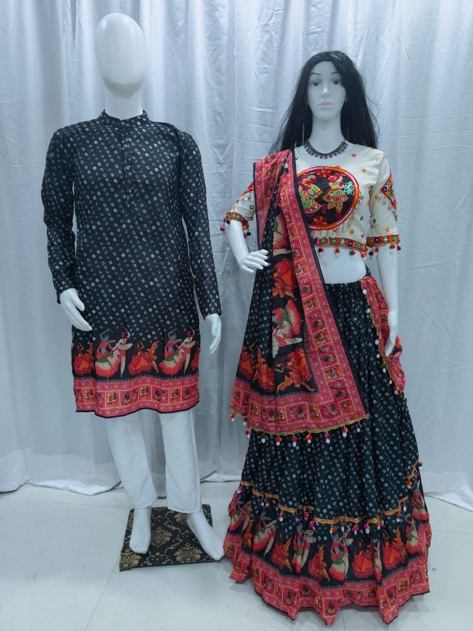 A traditional Indian ethnic Navratri Couple Wear Matching Outfit Set wear displayed on mannequins, featuring a black kurta with white prints and red border on the left, and a matching lehenga choli set on the right with a cream blouse, black skirt, and red patterned dupatta.