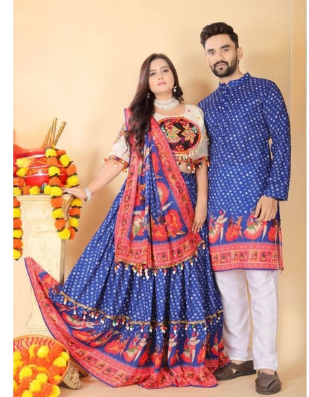 A couple modeling traditional Indian clothing, featuring a woman in a blue and red patterned lehenga choli with a matching dupatta, and a man in a matching blue kurta and white trousers, posed against a beige background with decorative marigold garlands.