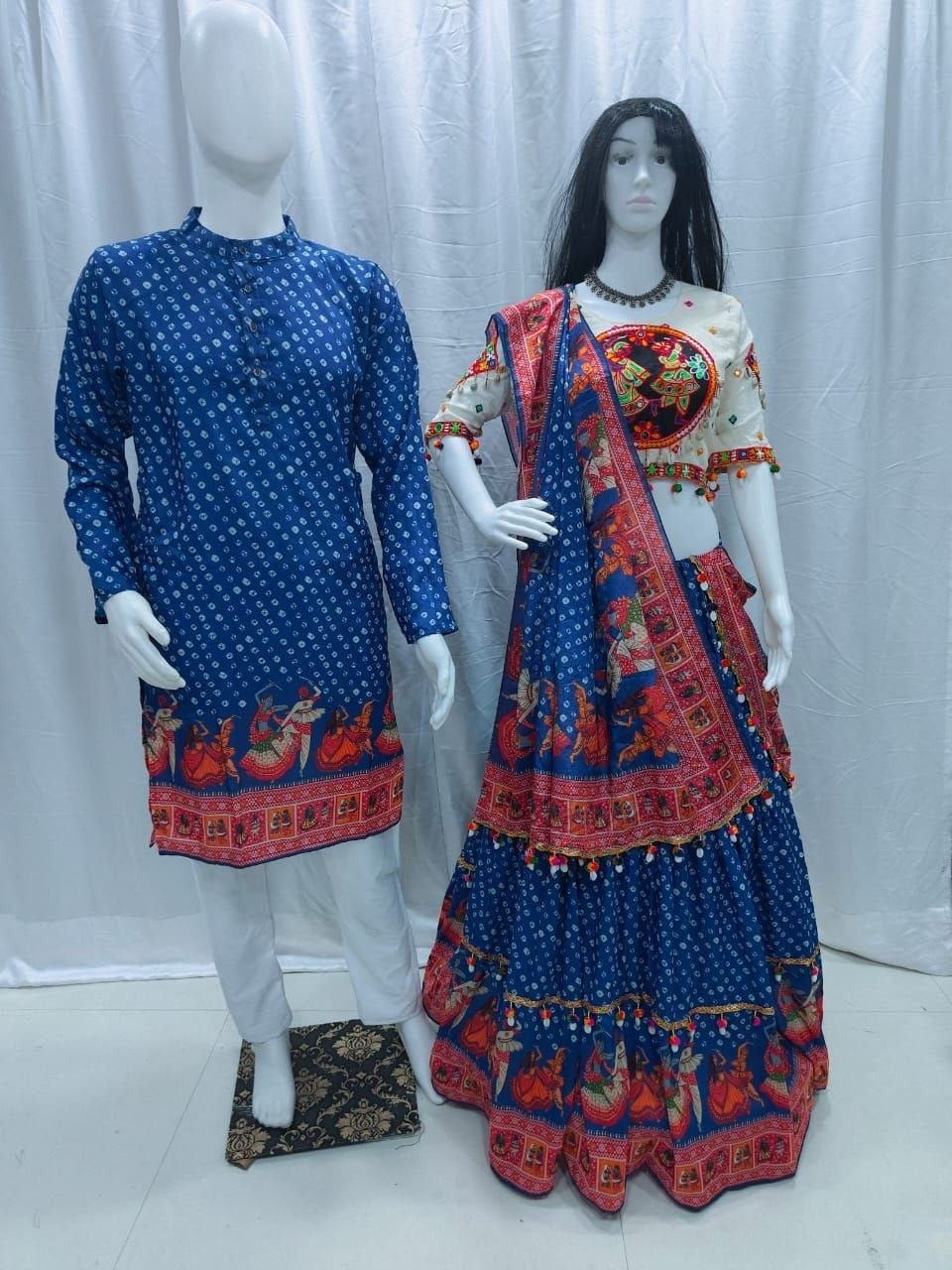 A traditional Indian outfit displayed on two mannequins against a white curtain backdrop. The mannequin on the left wears a blue kurta with white motifs and a red border print, paired with white trousers.