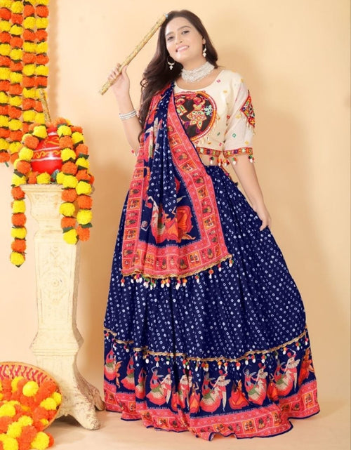 Load image into Gallery viewer, A woman modeling a traditional Indian ethnic outfit consisting of a navy blue flared skirt (lehenga) with white polka dots and red/orange border prints, paired with a cream-colored blouse featuring red and gold embroidery. She is holding a wooden stick and standing in front of a beige wall decorated with marigold garlands.
