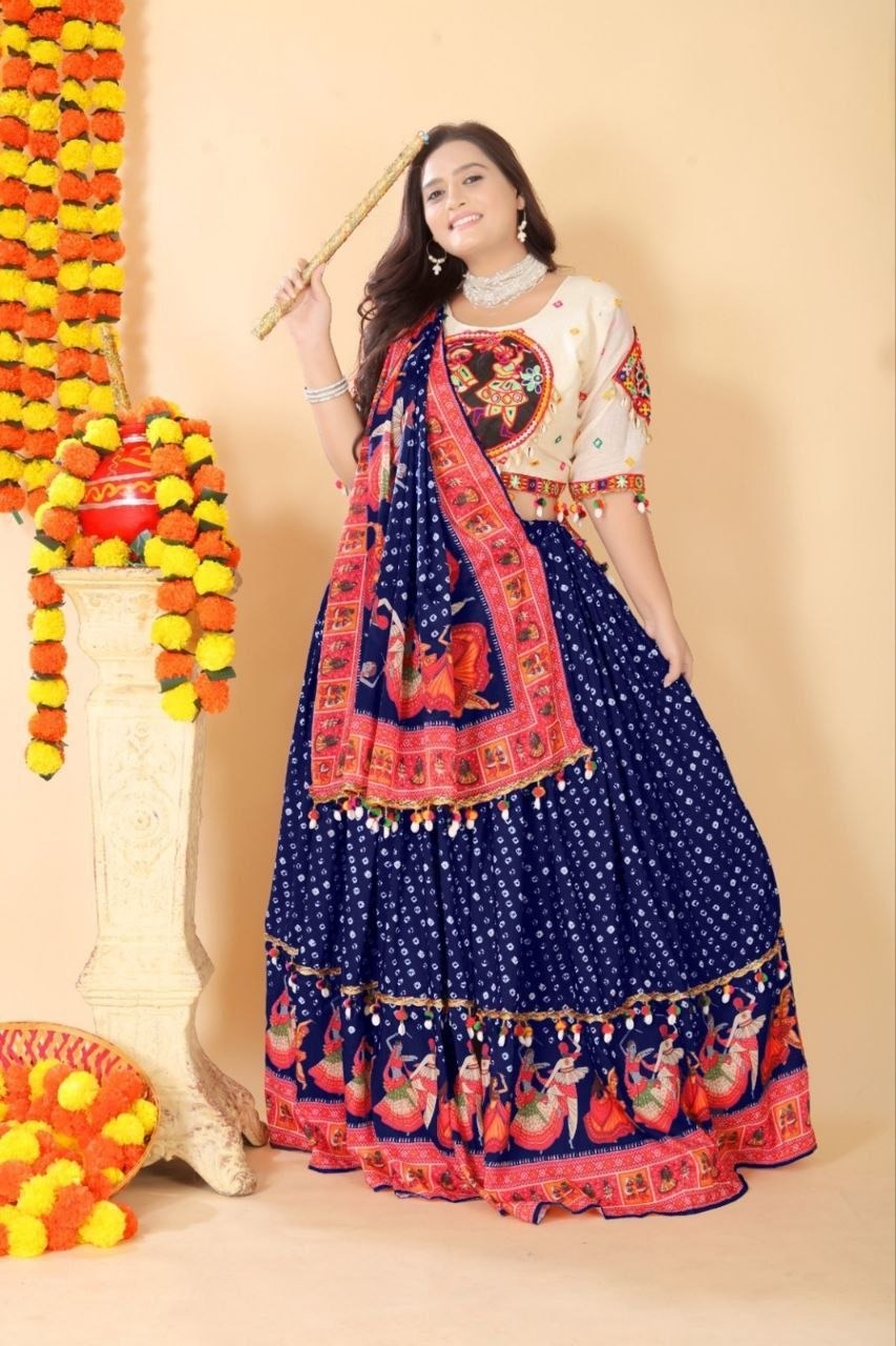 A woman modeling a traditional Indian ethnic outfit consisting of a navy blue flared skirt (lehenga) with white polka dots and red/orange border prints, paired with a cream-colored blouse featuring red and gold embroidery. She is holding a wooden stick and standing in front of a beige wall decorated with marigold garlands.