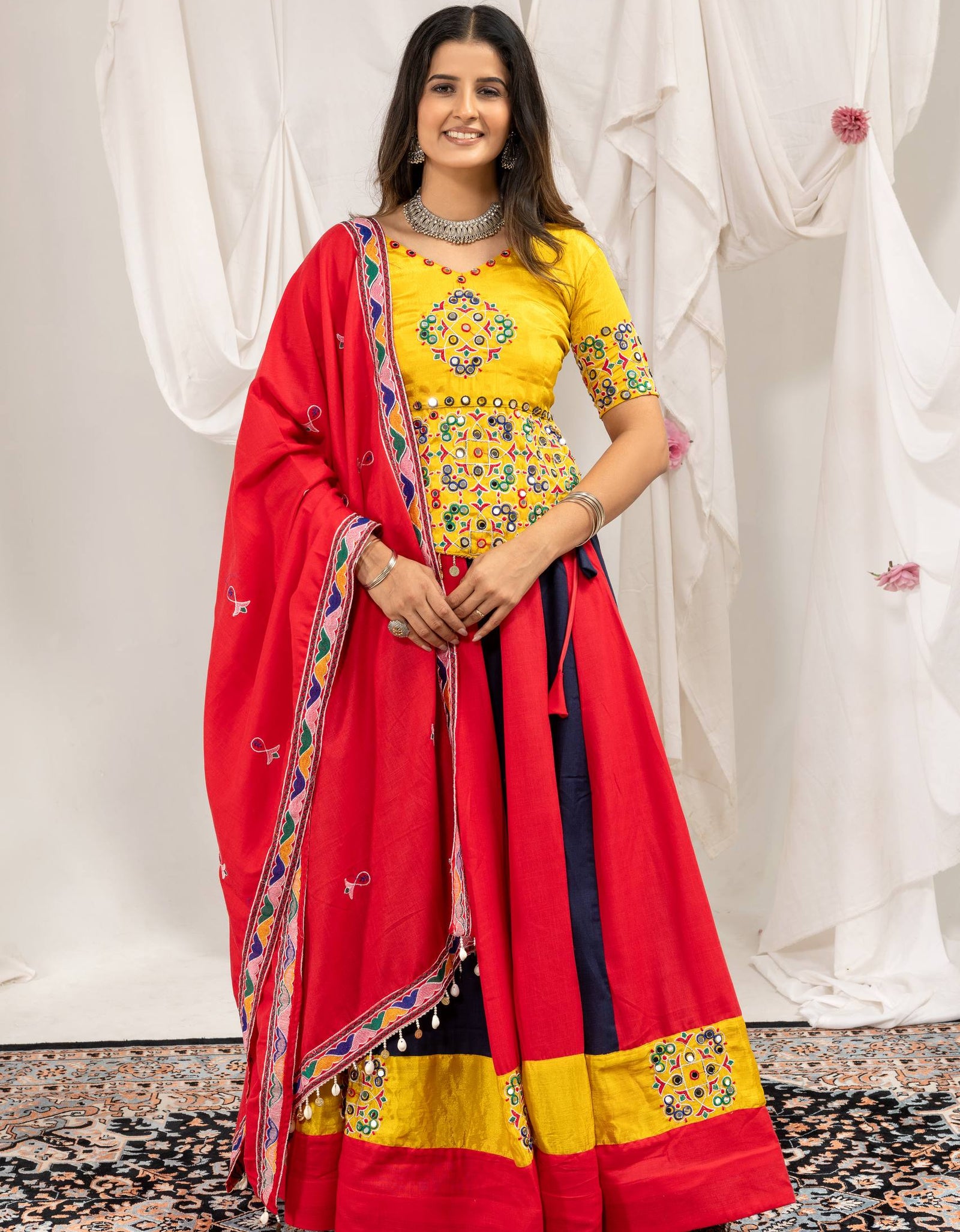 A woman modeling a traditional Indian lehenga choli set. The outfit features a bright yellow embroidered blouse with short sleeves, a matching red flared skirt (lehenga) with yellow and navy blue borders, and a red dupatta with a patterned border.