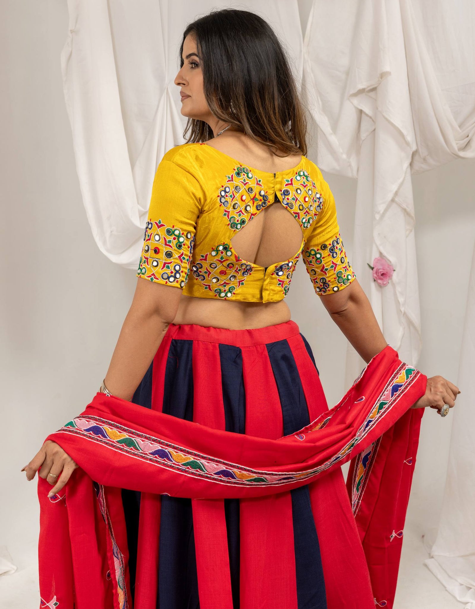 A woman modeling a traditional Indian outfit, shown from the back. She is wearing a bright yellow blouse with colorful floral embroidery and a back cutout, paired with a red and navy blue striped skirt and holding a red dupatta with a colorful border.