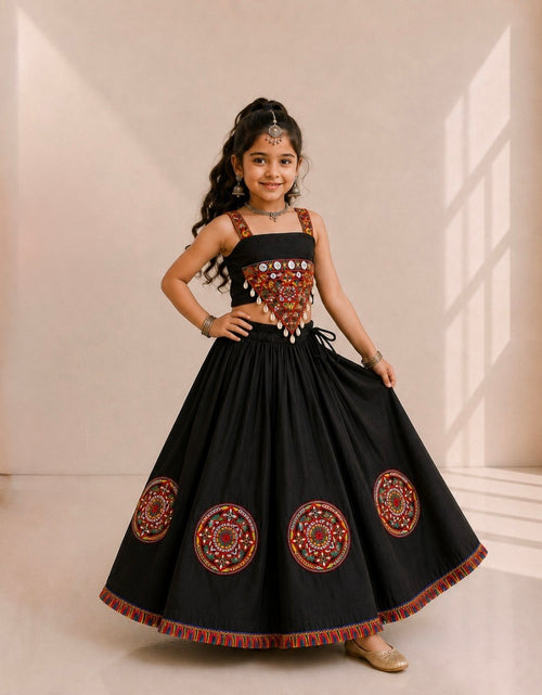 Load image into Gallery viewer, A young girl modeling a black traditional Indian dress with intricate red, gold, and silver embroidery on the bodice and circular patterns on the skirt, set against a warm beige background with window shadows.
