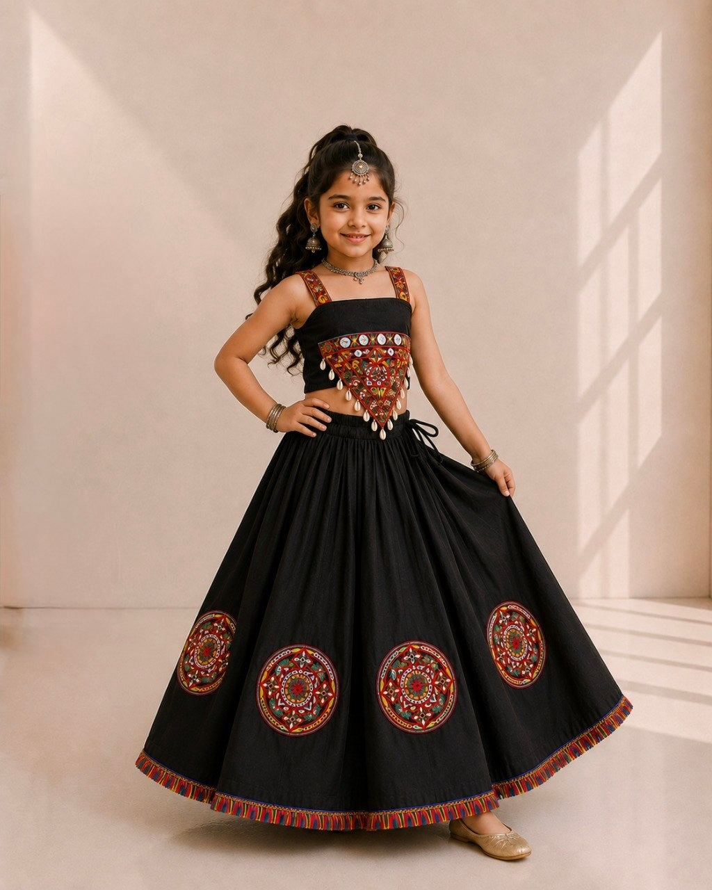 A young girl modeling a black traditional Indian dress with intricate red, gold, and silver embroidery on the bodice and circular patterns on the skirt, set against a warm beige background with window shadows.