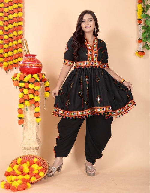 Load image into Gallery viewer, A woman modeling a black traditional Indian outfit with colorful embroidered details, paired with black pants, standing in front of a beige backdrop decorated with marigold garlands and a red pot.

