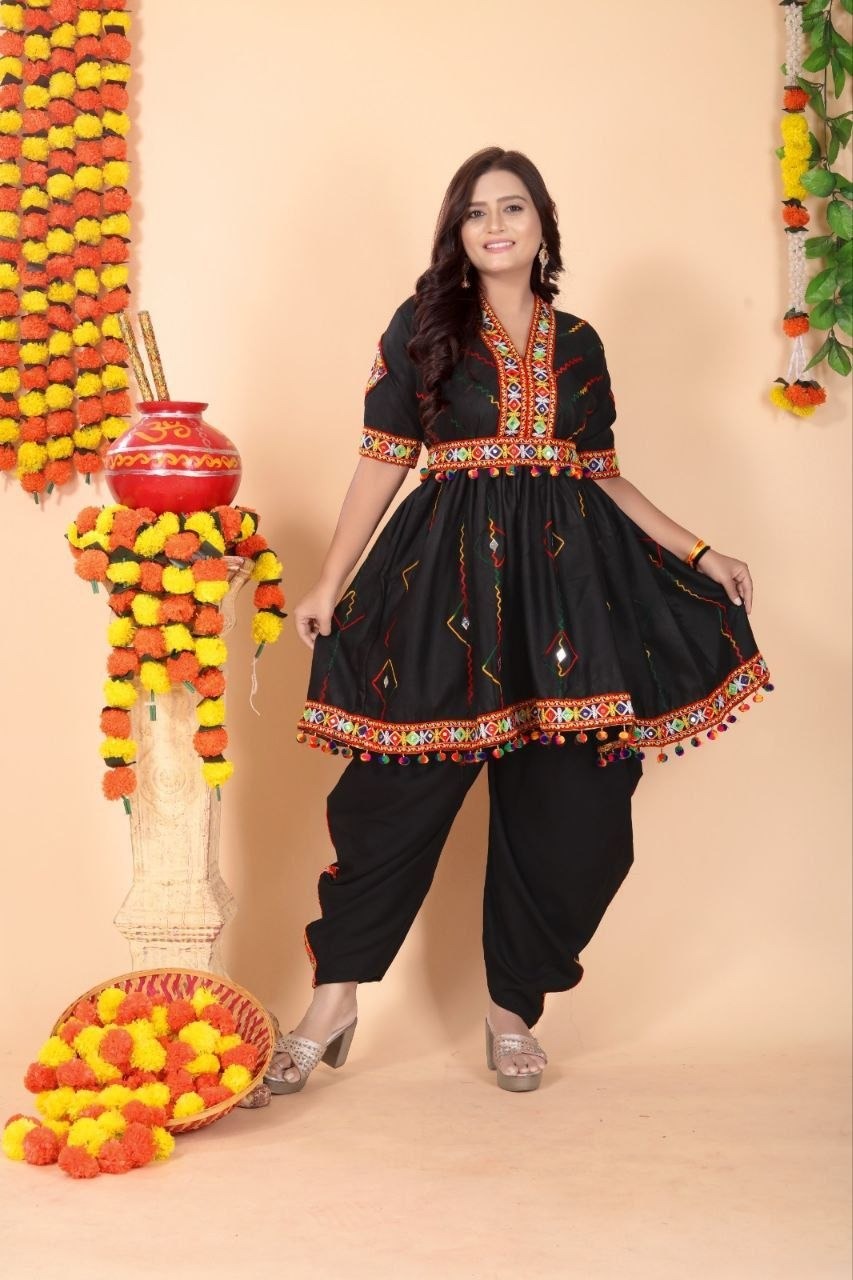 A woman modeling a black traditional Indian outfit with colorful embroidered details, paired with black pants, standing in front of a beige backdrop decorated with marigold garlands and a red pot.