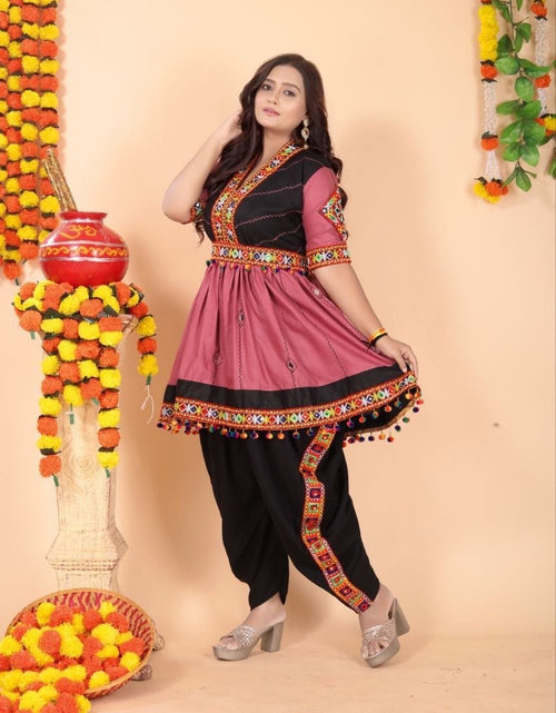 Load image into Gallery viewer, A woman modeling a traditional Indian salwar kameez outfit with a dusty pink tunic featuring black panels and colorful embroidered borders for Navratri dandiya nights, paired with black pants, against a peach-colored backdrop decorated with marigold garlands.

