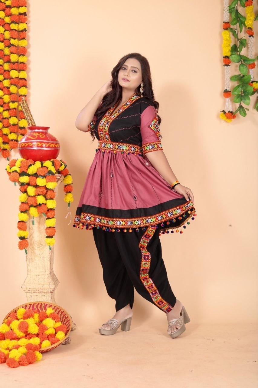 A woman modeling a traditional Indian salwar kameez outfit with a dusty pink tunic featuring black panels and colorful embroidered borders for Navratri dandiya nights, paired with black pants, against a peach-colored backdrop decorated with marigold garlands.