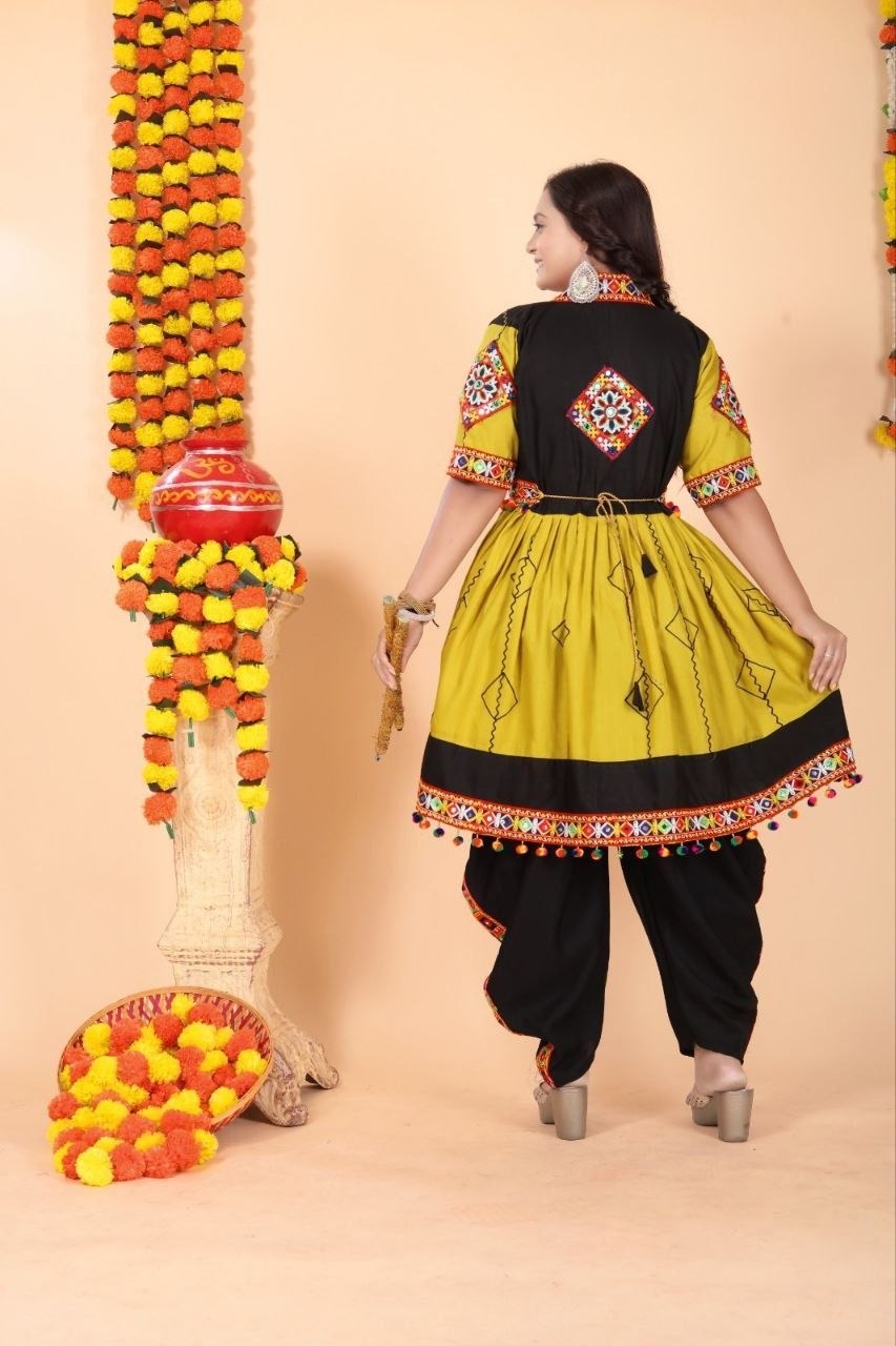 A woman modeling a traditional Indian salwar kameez outfit from the back, featuring a mustard yellow and black kurta with colorful embroidery, paired with black salwar pants, set against a beige background decorated with marigold garlands.