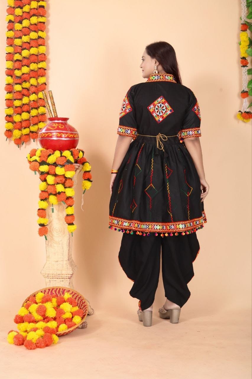 A woman wearing a black traditional Indian outfit with colorful embroidered details on the sleeves, hem, and back, standing in front of a beige backdrop decorated with marigold garlands and a red pot.