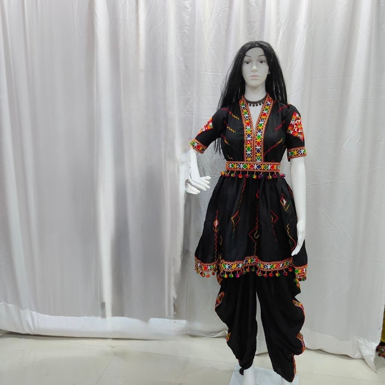 A black traditional outfit displayed on a mannequin against a white curtain backdrop. The outfit includes a short-sleeve top with colorful embroidered details around the neckline and sleeves, a matching embroidered waist tie, and black pants with decorative trim.