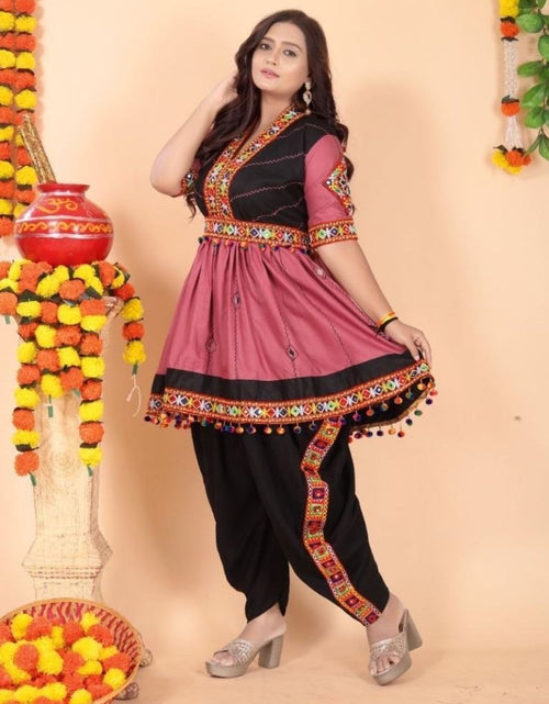 Load image into Gallery viewer, A woman models a traditional Indian Navratri salwar kameez outfit with a dusty pink tunic, black pants, and a black collar panel, decorated with colorful embroidered borders and pom-pom accents. The background is a warm beige studio setting decorated with marigold garlands.
