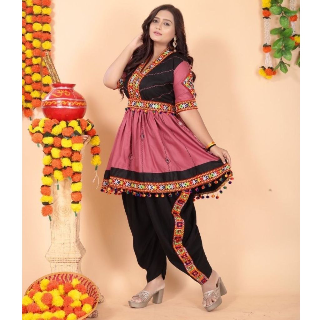 A woman models a traditional Indian Navratri salwar kameez outfit with a dusty pink tunic, black pants, and a black collar panel, decorated with colorful embroidered borders and pom-pom accents. The background is a warm beige studio setting decorated with marigold garlands.
