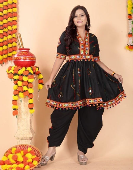 A woman modeling a black traditional Indian outfit with colorful embroidered details, featuring a kurta with a flared skirt and matching black pants, posed against a peach-colored backdrop decorated with marigold garlands.