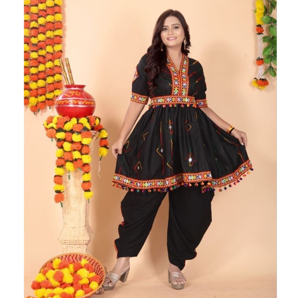 A woman modeling a black traditional Indian outfit with colorful embroidered details, featuring a kurta with a flared skirt and matching black pants, posed against a peach-colored backdrop decorated with marigold garlands.