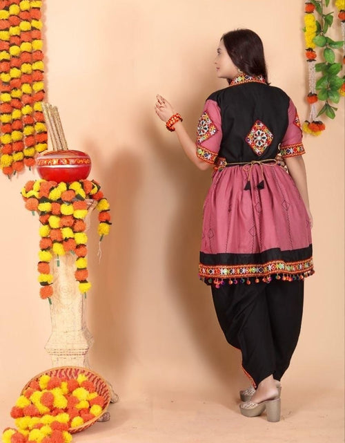 Load image into Gallery viewer, A woman wearing a traditional Indian Navratri outfit with a pink and black kurta, black pants, and silver heels, standing in front of a beige wall decorated with marigold garlands.
