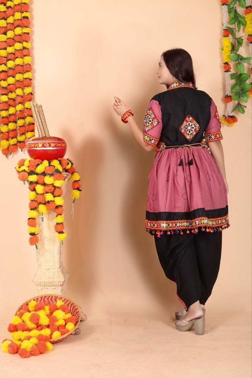 A woman wearing a traditional Indian Navratri outfit with a pink and black kurta, black pants, and silver heels, standing in front of a beige wall decorated with marigold garlands.