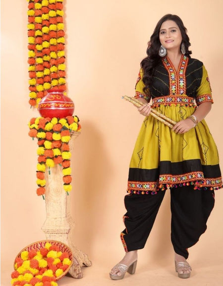 A woman models a traditional Indian outfit consisting of a mustard yellow and black kurta with colorful embroidery, paired with black pants. She is standing next to a decorative garland stand against a solid beige background.