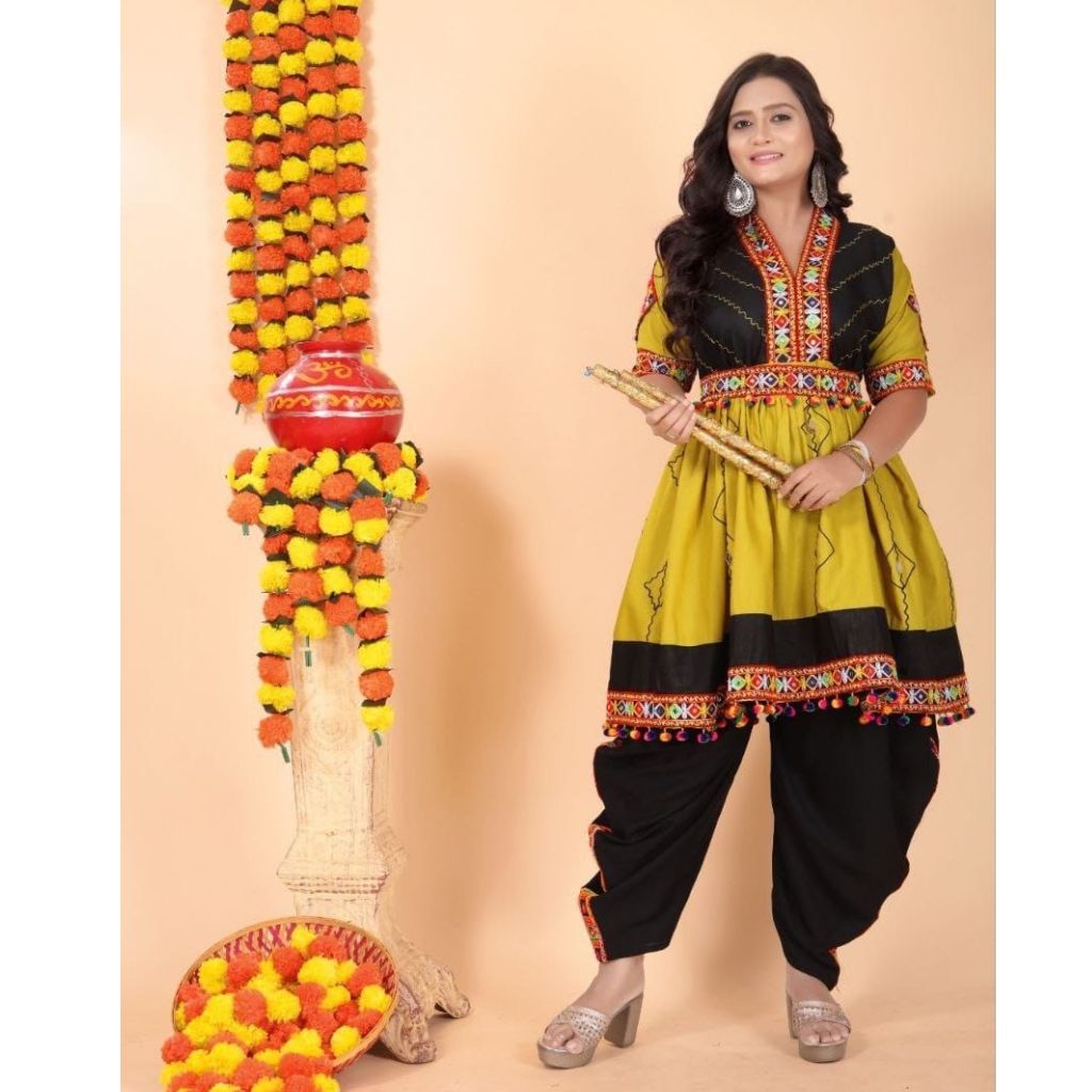 A woman models a traditional Indian outfit consisting of a mustard yellow and black kurta with colorful embroidery, paired with black pants. She is standing next to a decorative garland stand against a solid beige background.